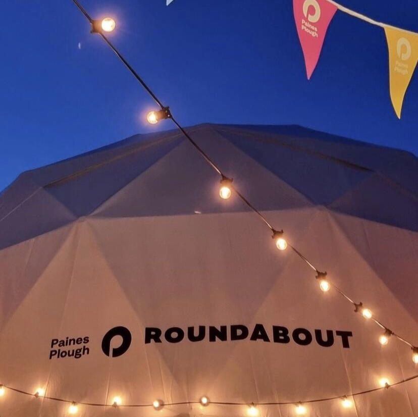 The outside of of a white exterior stage-like dome that says Roundabout while strings lights light up the exterior.