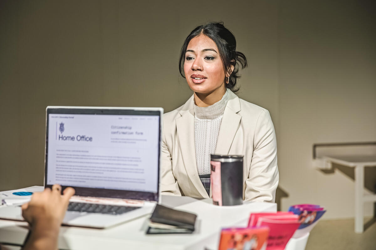 An actor sitting in front of a desk wearing a white professional outfit and makeup while a person is on a laptop in front of them.