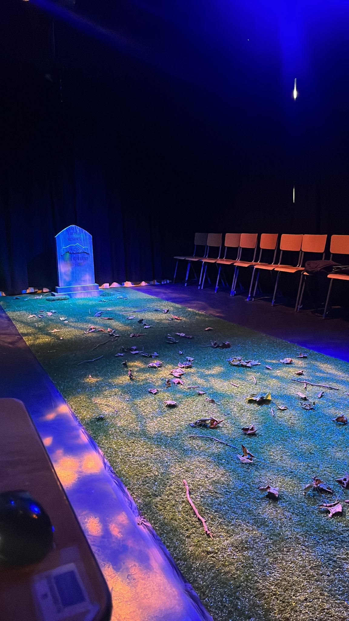 A stage with a tombstone and leaves on grass while chairs are lined up around it.